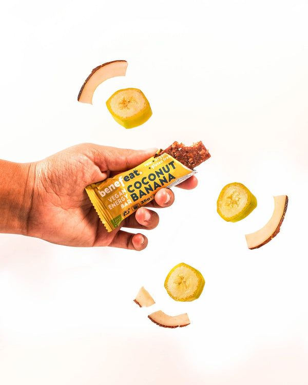 Hand holding an open Coconut Banana bar with coconut and banana slices flying around